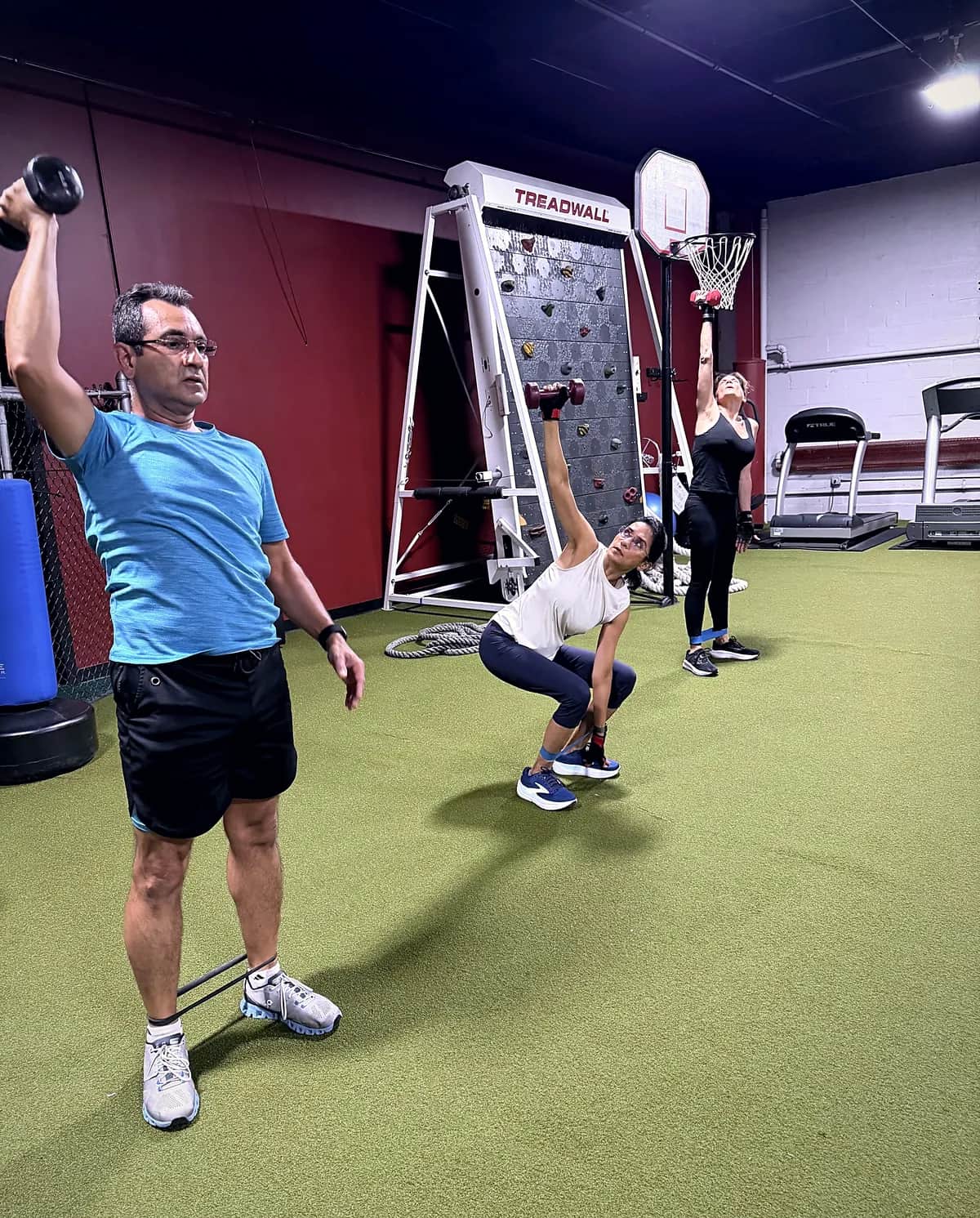 Three people training with dumbbells and a resistance band on the turf floor, a climbing wall behind them